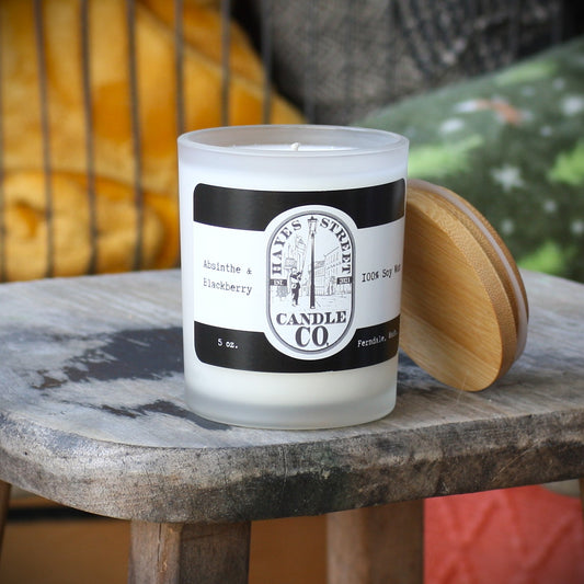 A frosted glass candle jar labeled 'Hayes Street Candle Co. Absinthe & Blackberry' sits on a rustic wooden table with its light wood lid resting beside it.