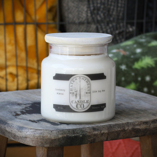 A large white soy wax candle in a clear glass jar with a light wood lid, sitting on a rustic wooden stool. The label reads 'Hayes Street CANDLE CO' and 'Cranberry Mimosa'. 18 oz Ferndale, Wash