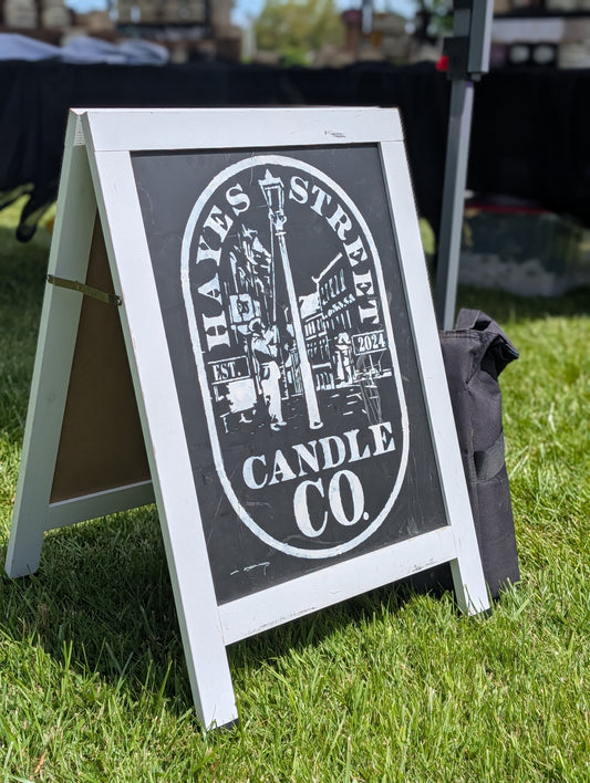 Hayes Street Candle Company sign set up on green grass. The logo is white, the background is black chalkboard, and the frame is white. It is a sunny day. Hayes Street Candle Company products are hand-poured by Margie Creen in Ferndale, Washington.