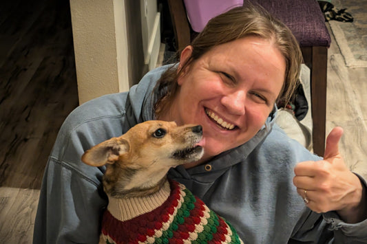 Margie Creen of Hayes Street Candle Company holding Chippy the dog. She is smiling and giving a thumbs up. Chippy is wearing a green, red, and cream holiday striped sweater and is licking her face.