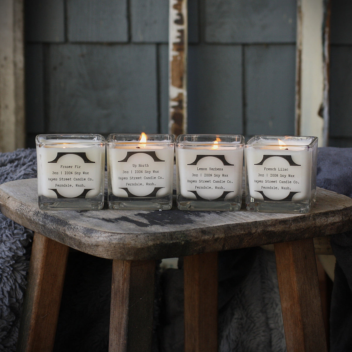 Four lit soy candles in clear square glass jars arranged on a rustic wooden stool, with a dark gray shingled wall in the background.