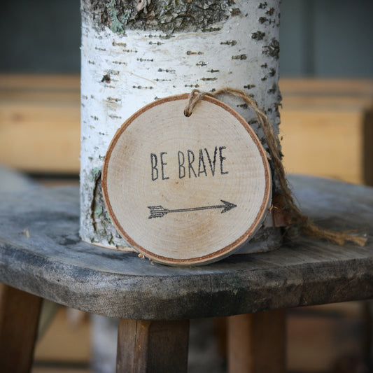 A round natural wood slice ornament hanging from twine, printed with the words 'BE BRAVE' and an arrow, displayed against a birch tree trunk on a rustic wooden stool. Made by Log Slices by Margie