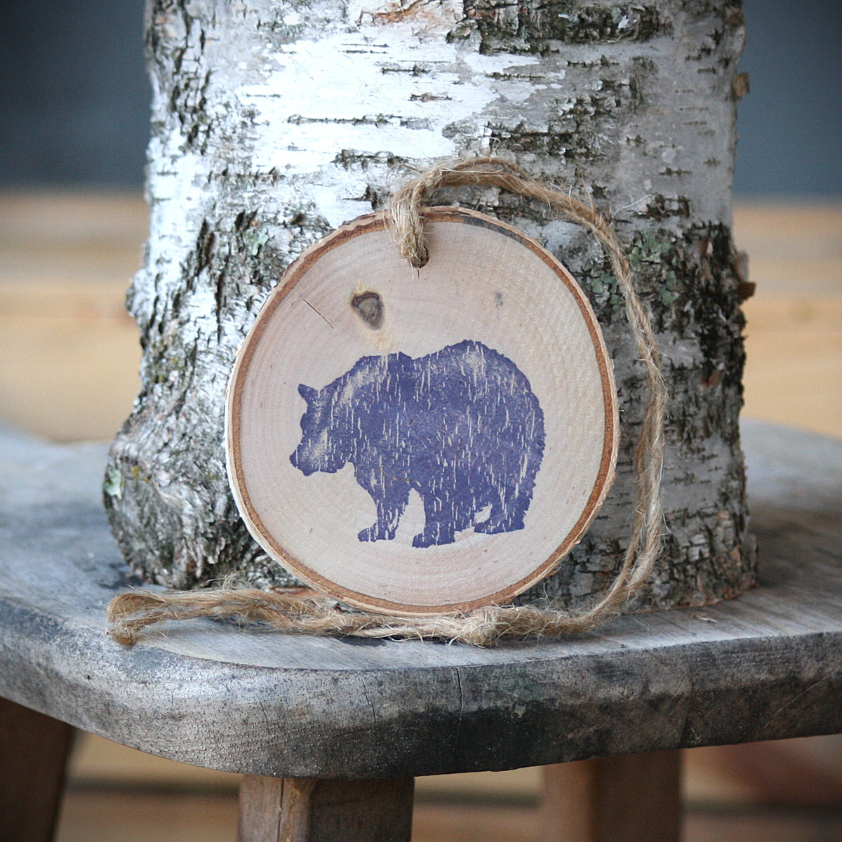 A round wooden slice ornament with a blue bear silhouette, hanging from twine on a rustic wooden stool in front of a birch tree trunk.