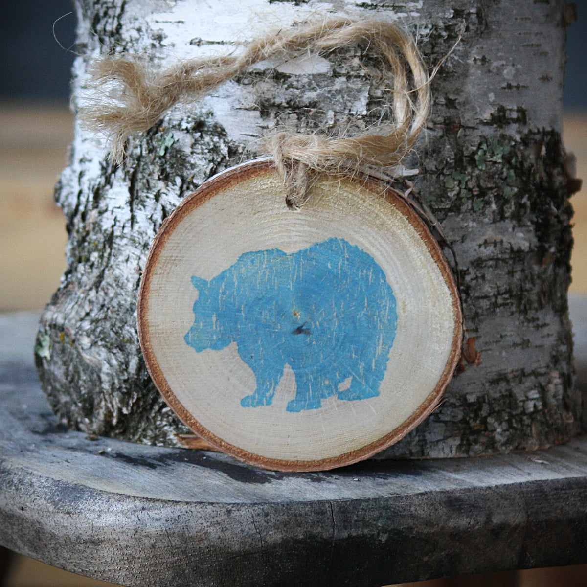 A round wooden slice ornament with a light blue bear silhouette, hanging from natural jute twine against a birch tree trunk. Made by Log Slices by Margie and Hayes Street Candle Company
