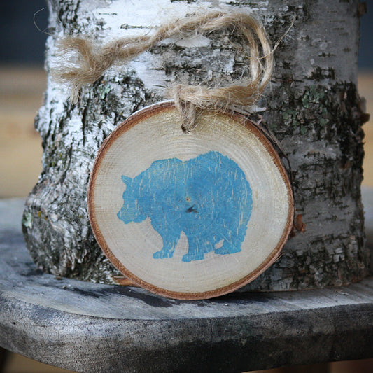 A round wooden slice ornament with a light blue bear silhouette, hanging from natural jute twine against a birch tree trunk. Made by Log Slices by Margie and Hayes Street Candle Company