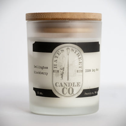 A frosted glass jar candle with a light wood lid, featuring a white wax fill and a black-and-white label for Hayes Street Candle Co. in the scent name 'Bellingham Blackberry'.