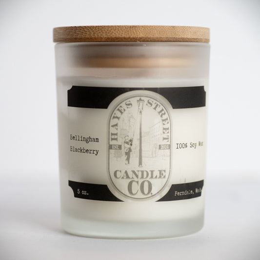 A frosted glass jar candle with a light wood lid, featuring a white wax fill and a black-and-white label for Hayes Street Candle Co. in the scent name 'Bellingham Blackberry'.