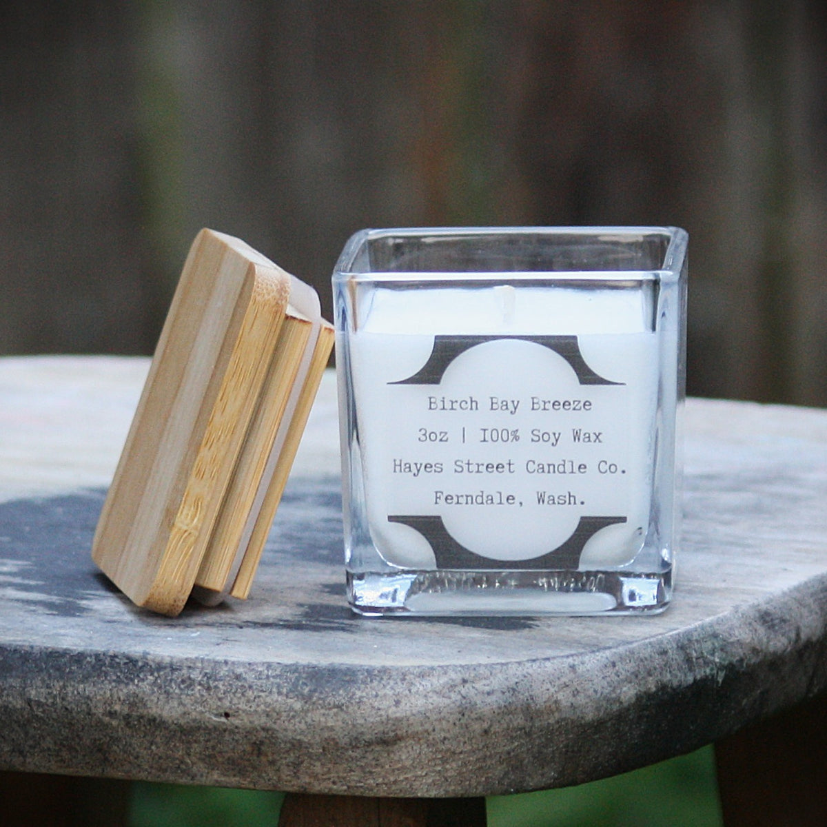 A square glass jar candle labeled 'Birch Bay Breeze' by Hayes Street Candle Co. sits on a rustic wooden table outdoors, with its bamboo lid leaning against it.