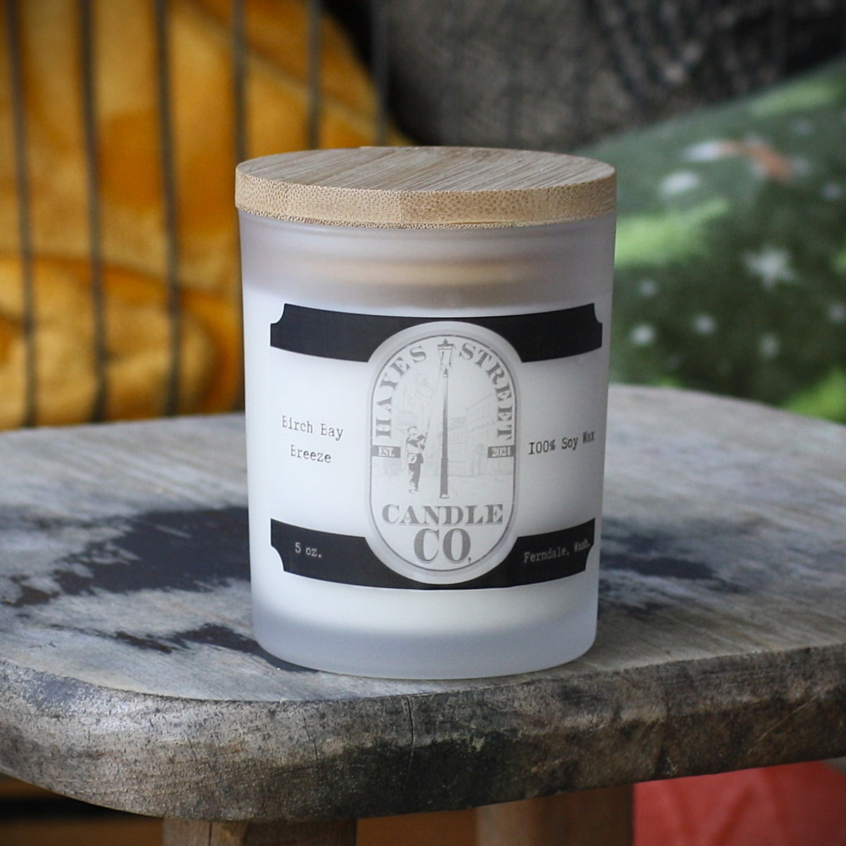 A frosted glass candle jar with a light wood lid sits on a rustic stone table. The front label reads Hayes Street Candle Co. With a scent of Birch Bay Breeze 5oz Ferndale Wash