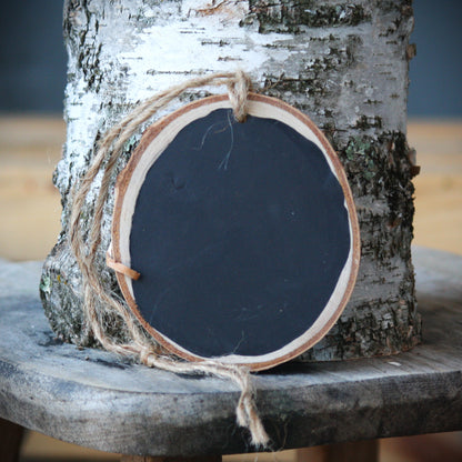 A round wooden sign with a black chalkboard center, natural wood edges, and a piece of jute twine hanging from the top, displayed against a birch tree trunk on a rustic wooden surface.