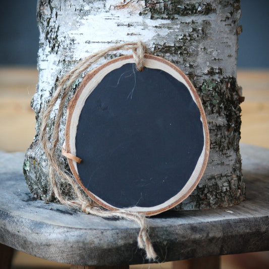 A round wooden sign with a black chalkboard center, natural wood edges, and a piece of jute twine hanging from the top, displayed against a birch tree trunk on a rustic wooden surface.
