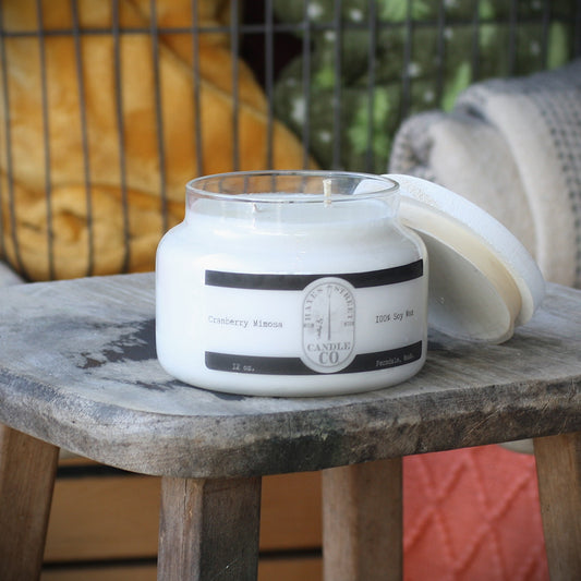 A white soy wax candle in a clear glass jar sits on a rustic wooden stool, with its lid leaning against the right side. The label reads Hayes Street Candle Company and Cranberry Mimosa