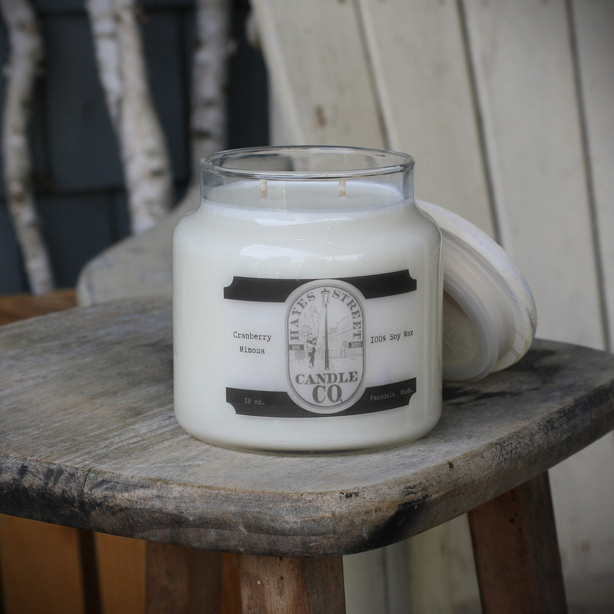 A large white soy wax candle in a clear glass jar sits on a rustic wooden stool, with its white lid leaning against the stool beside it. The label reads 'HAYES STREET CANDLE CO' and 'Cranberry Mimosa'