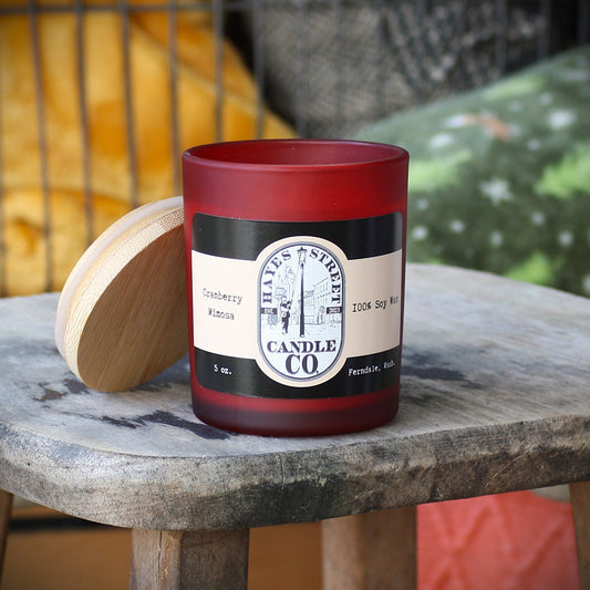 A red glass candle jar with a cream and black label reading Hayes Street Candle Co., placed on a rustic wooden table with its wooden lid beside it. Cranberry Mimosa