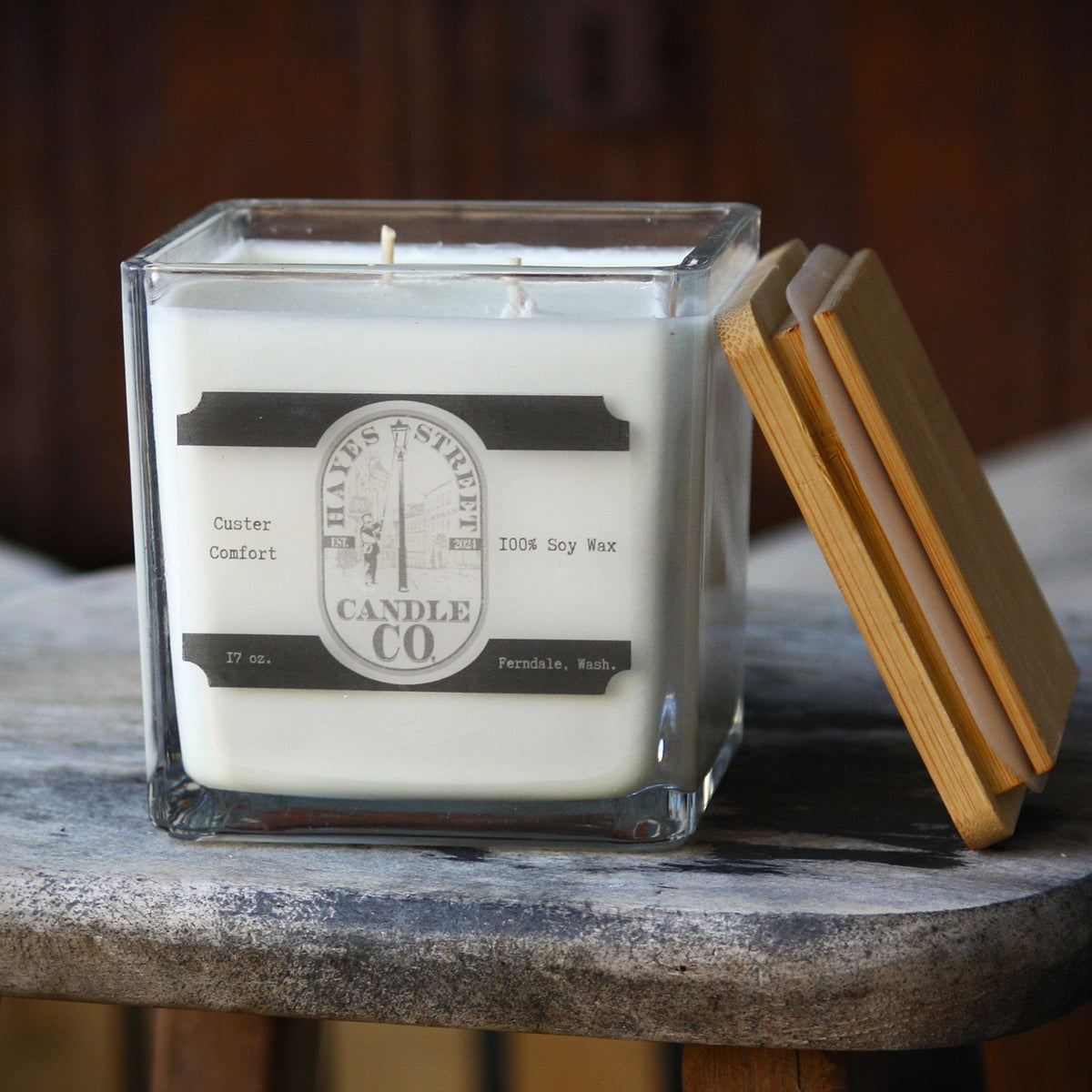 A square glass jar candle with white wax sits on a rustic wood surface, with a bamboo lid leaning against the right side. The front label reads Hayes Street Candle Co.