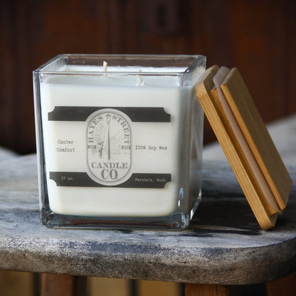 A square glass jar candle with white wax sits on a rustic wood surface, with a bamboo lid leaning against the right side. The front label reads Hayes Street Candle Co.