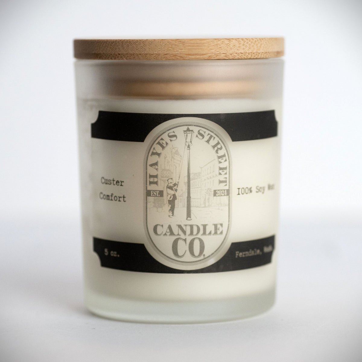 A 5 oz soy wax candle in a frosted glass jar with a light wood lid, featuring a cream and black label for Hayes Street Candle Co. The scent is pumpkin, caramel, and cream, and is called Custer Comfort. Ferndale, Wash