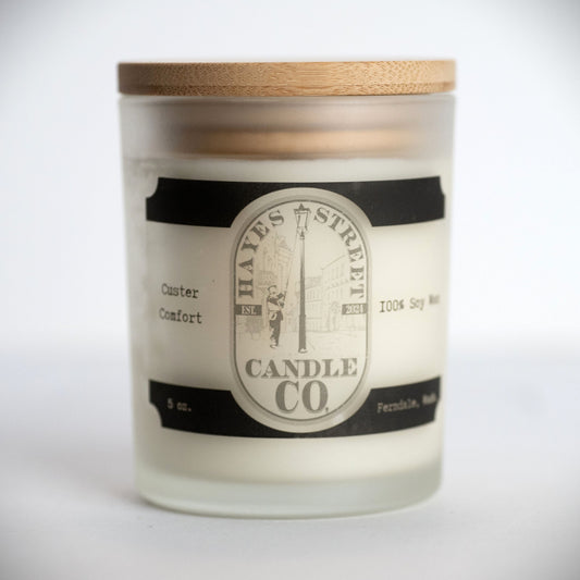 A 5 oz soy wax candle in a frosted glass jar with a light wood lid, featuring a cream and black label for Hayes Street Candle Co. The scent is pumpkin, caramel, and cream, and is called Custer Comfort. Ferndale, Wash
