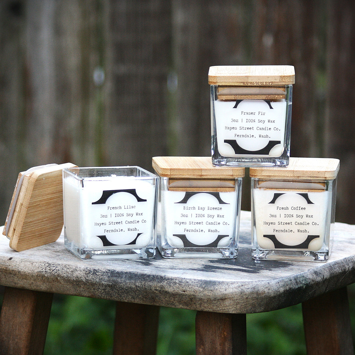 Four square glass jar candles with bamboo lids are arranged on a rustic wood stool outdoors, with one lid leaning beside them. The candles have white wax and black-and-white labels reading 'French Lilac,' 'Birch Bay Breeze,' "Fraser Fir,' and 'Fresh Coffee.' They are made by Hayes Street Candle Company