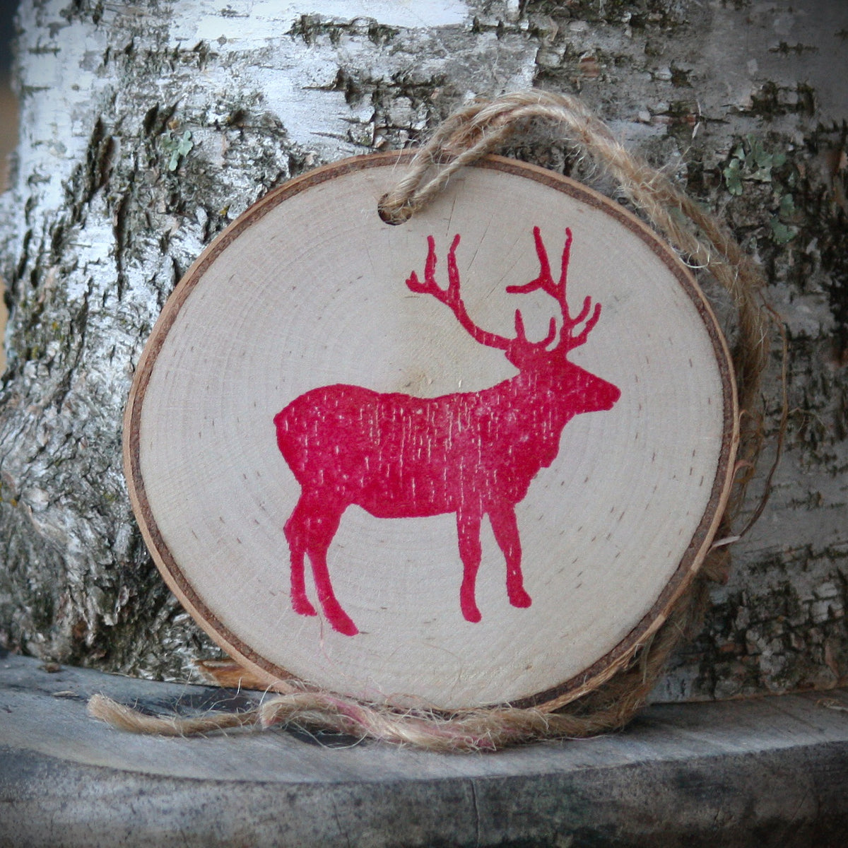 Elk silhouette in red stamped ink on paper birch log slice ornament, leaning against a birch stump on a rustic wooden stool. Made by Log Slices by Margie and sold by Hayes Street Candle Company.