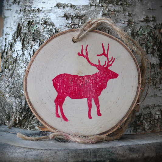 Elk silhouette in red stamped ink on paper birch log slice ornament, leaning against a birch stump on a rustic wooden stool. Made by Log Slices by Margie and sold by Hayes Street Candle Company.