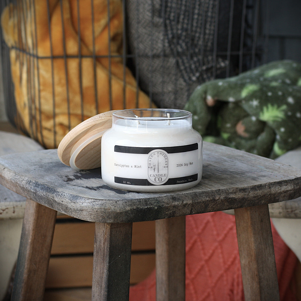 A white soy wax candle in a clear glass jar with a light wood lid resting beside it on a rustic wooden stool. The label reads Hayes Street Candle Company