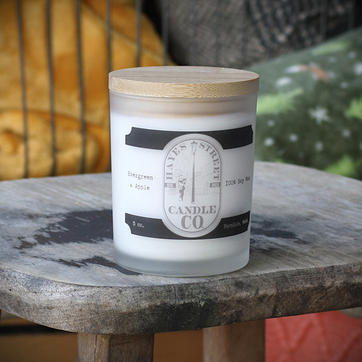 Evergreen and Apple 5oz glass soy wax candle with Hayes Street Candle Company on the logo. The candle has a bamboo lid and is resting on a worn wooden stool.