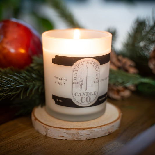 A lit white candle in a frosted glass jar sits on a round wood slice coaster. The candle label reads 'Hayes Street Candle Co' and 'Evergreen & Apple'.