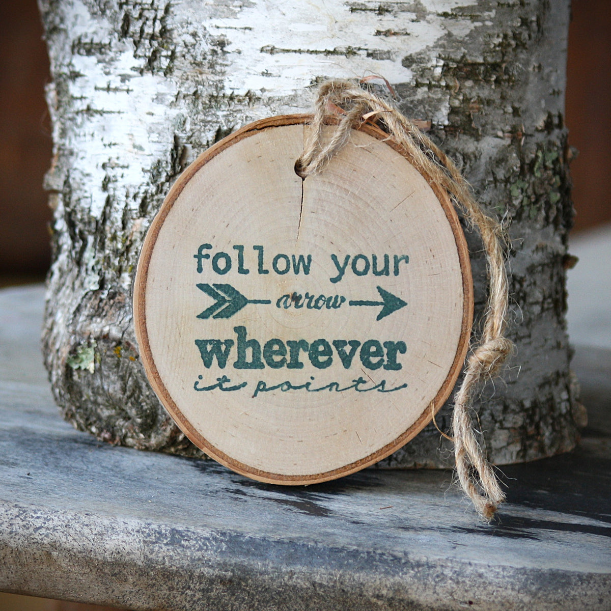 Follow our arrow wherever it points log slice ornament made of birch wood. In the photo it is leaning against a piece of white birch wood bark on a rustic wooded stool, made by Log Slices by Margie and Hayes Street Candle Company