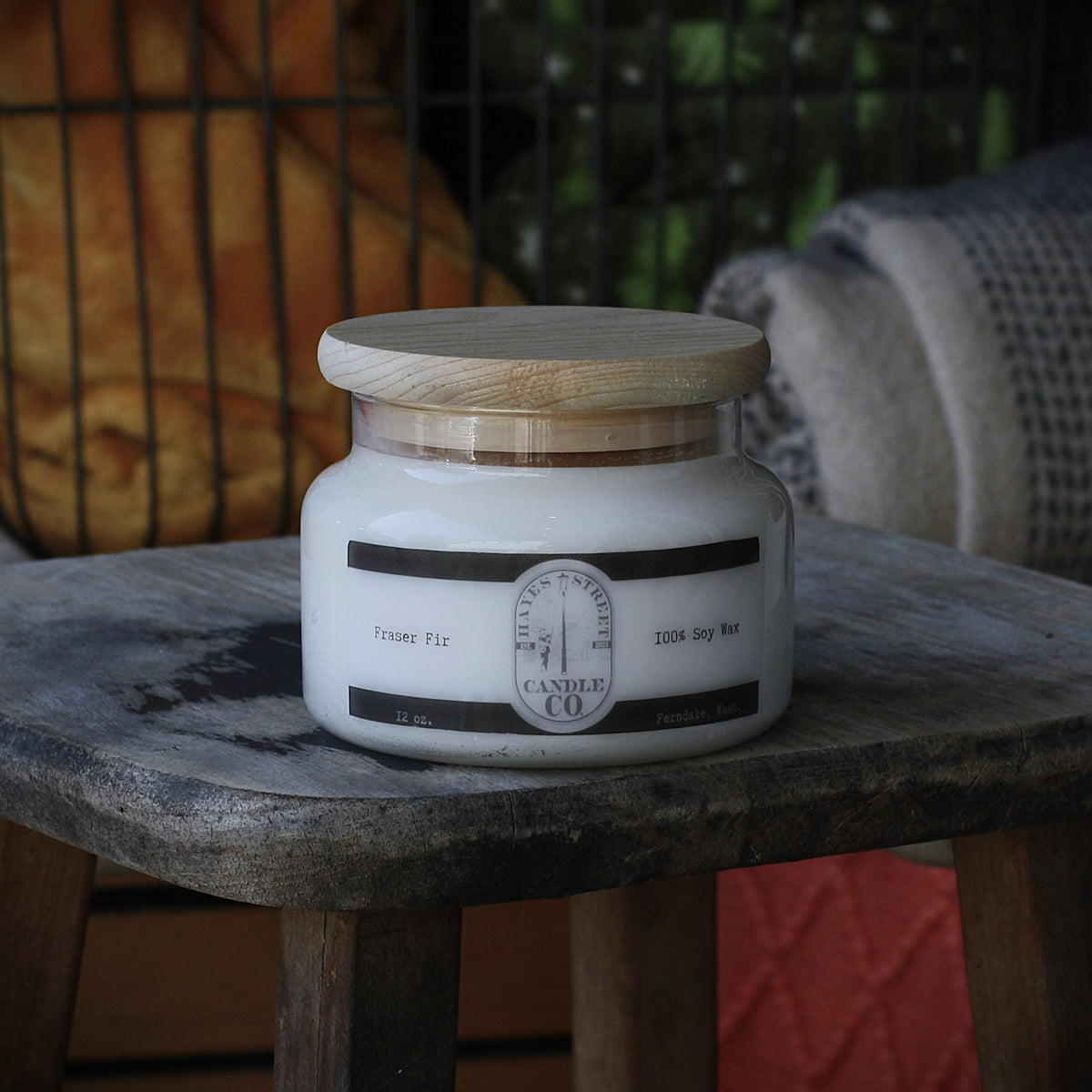 A white soy wax candle in a clear glass jar with a light wood lid, sitting on a rustic wooden stool. The label reads Hayes Street Candle Company. Fraser Fir