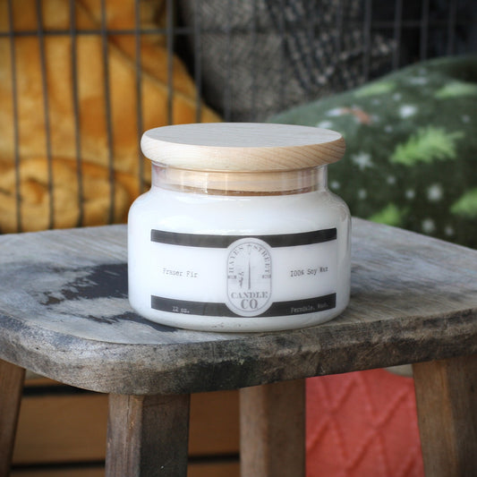 A white candle in a clear glass jar with a light wood lid, sitting on a rustic wooden stool. The label reads 'Hayes Street Candle Co' and Fraser Fir. 100% Soy Wax and Ferndale, Wash.