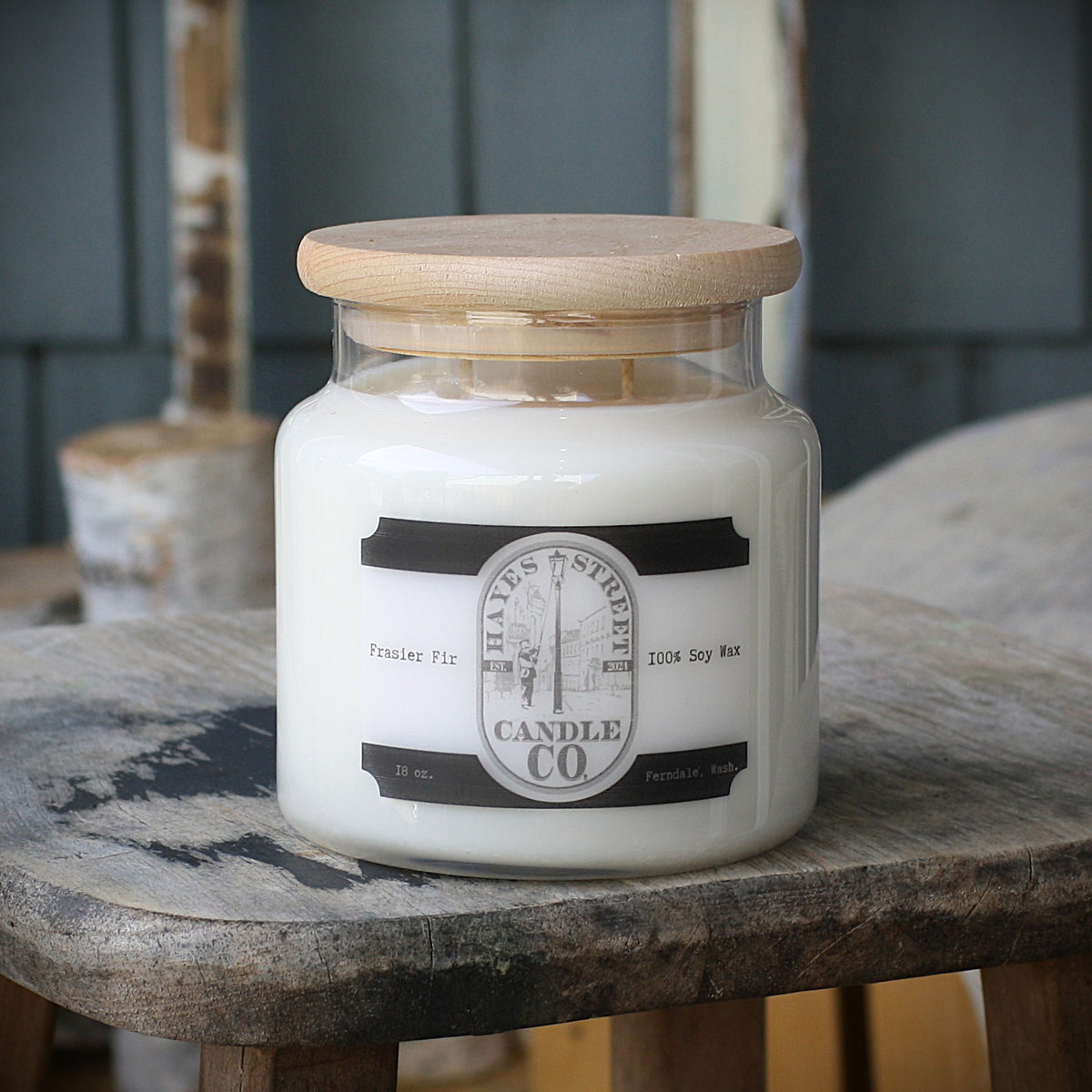 An 18 oz soy wax candle in a clear glass jar with a light wood lid, resting on a rustic wooden stool. The label reads 'HAYES STREET CANDLE CO' and 'Fraser Fir'.