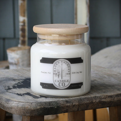 An 18 oz soy wax candle in a clear glass jar with a light wood lid, resting on a rustic wooden stool. The label reads 'HAYES STREET CANDLE CO' and 'Fraser Fir'.