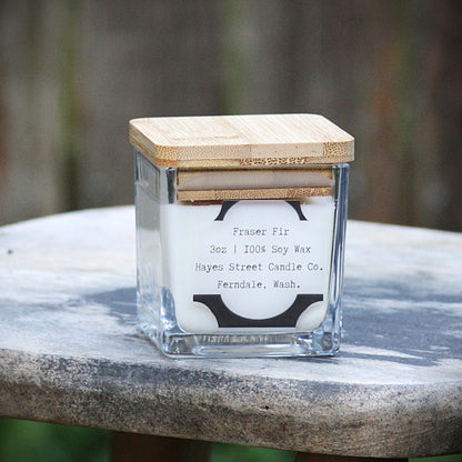 Small square candle made of glass with a bamboo lid resting on a rustic wooden stool. The label reads Fraser Fir 3oz 100% soy wax Hayes Street Candle Co. Ferndale, Wash.