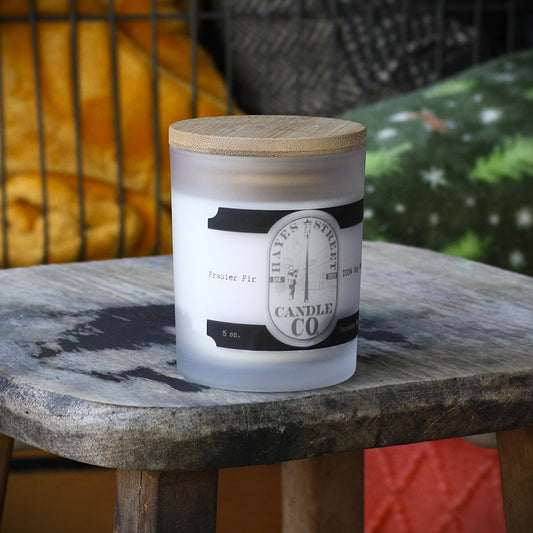 A frosted glass candle jar with a light wood lid sits on a rustic wooden stool. The label reads  Hayes Street Candle Company and Ferndale Wash
