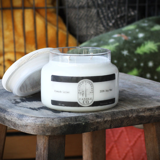 A large white soy wax candle in a clear glass jar with a white lid leaning beside it, placed on a rustic wooden stool. The label reads 'French Lilac' and 'Hayes Street Candle Co.