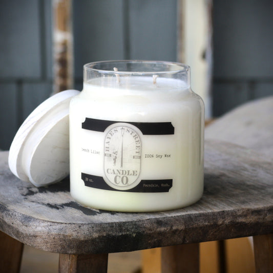 A large white soy wax candle in a clear glass jar sits on a rustic wooden stool, with its white lid resting beside it. The label reads 'HAYES STREET CANDLE CO' and 'Fresh Lilac'.