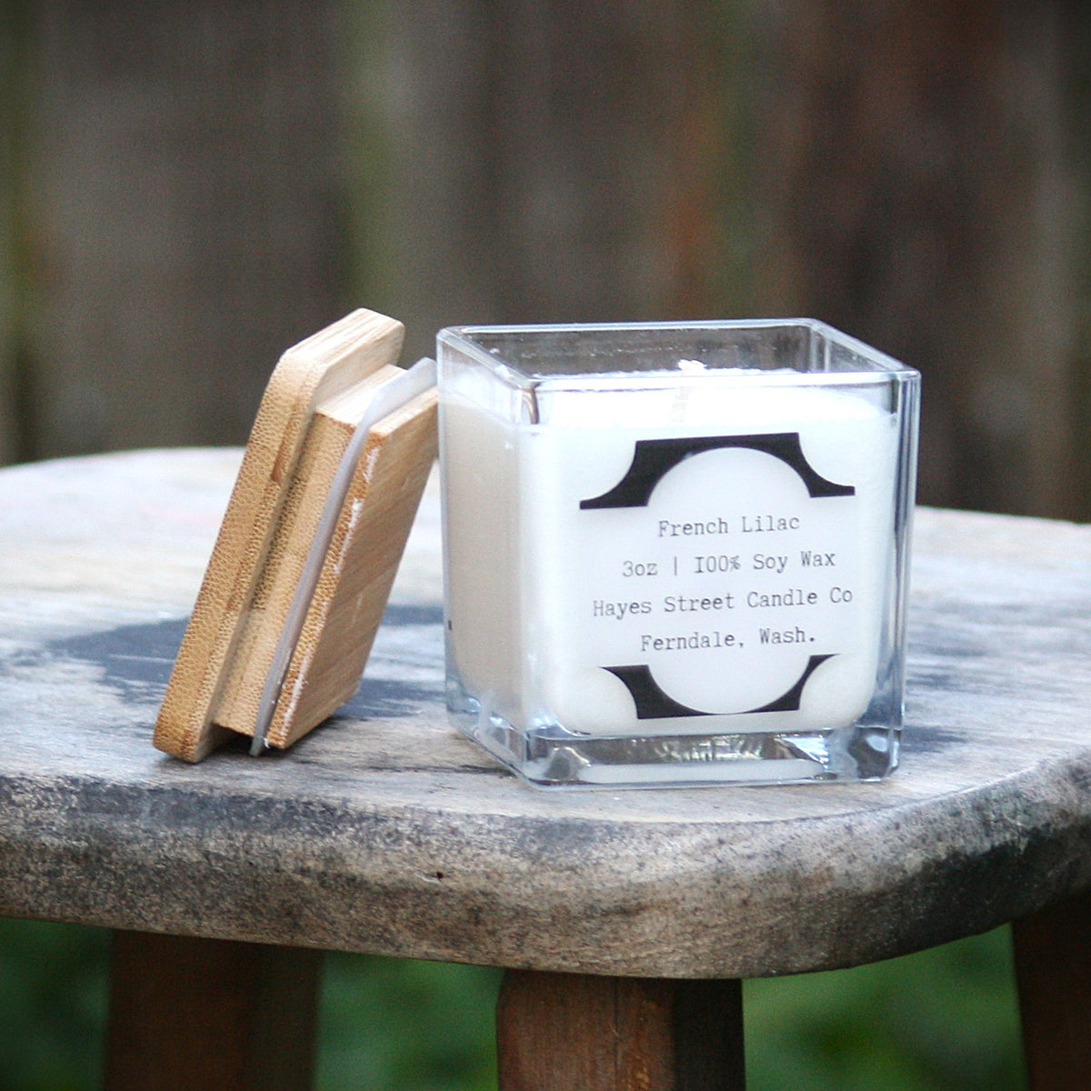 Small square candle jar with French Lilac on it. It is resting on a distressed vintage wood stool, with a blurred out rustic fence behind it, and a little of blurred green grass visible. The label also reads 3oz 100% soy wax Hayes Street Candle Co Ferndale, Wash.
