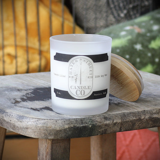 French Lilac candle in a straight-sided glass jar. The bamboo lid is set on the side and is leaning against the glass. It is resting on a rustic wooden stool with colorful blankets behind it.