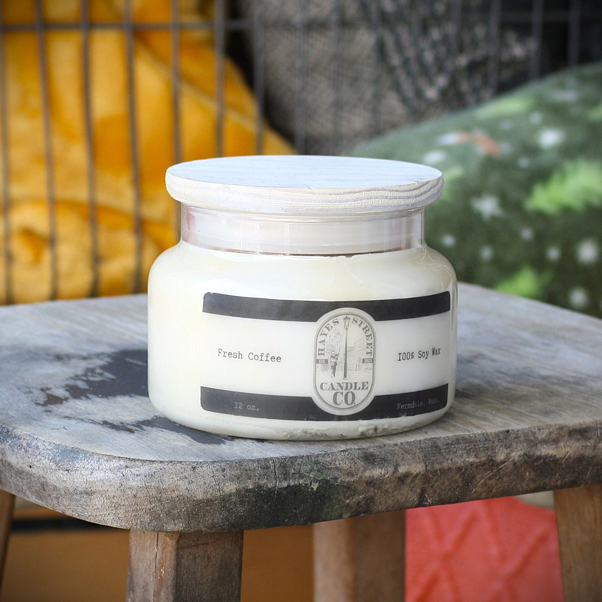 A 12 oz glass jar candle with a white wax fill and a white lid, featuring a black and white label that reads 'Fresh Coffee' and 'Hayes Street Candle Co'. The candle is displayed on a rustic wooden stool with a blurred background containing yellow and green cushions.