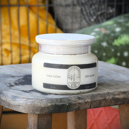 A 12 oz glass jar candle with a white wax fill and a white lid, featuring a black and white label that reads 'Fresh Coffee' and 'Hayes Street Candle Co'. The candle is displayed on a rustic wooden stool with a blurred background containing yellow and green cushions.