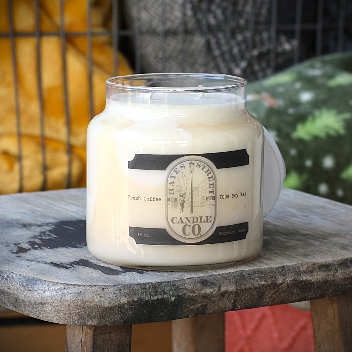 A large glass jar candle labeled 'Hayes Street Candle Co. French Coffee' sits on a rustic wooden stool in a cozy indoor setting with a yellow blanket and patterned pillows in the background.