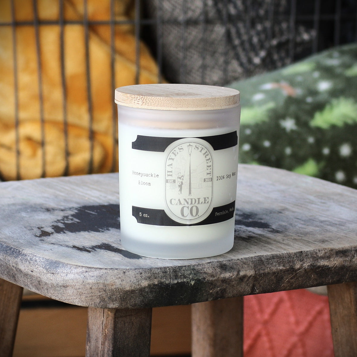 A frosted glass jar candle with a bamboo lid sits on a weathered wooden table. The label reads Hayes Street Candle Company. The scent is Honeysuckle Bloom.