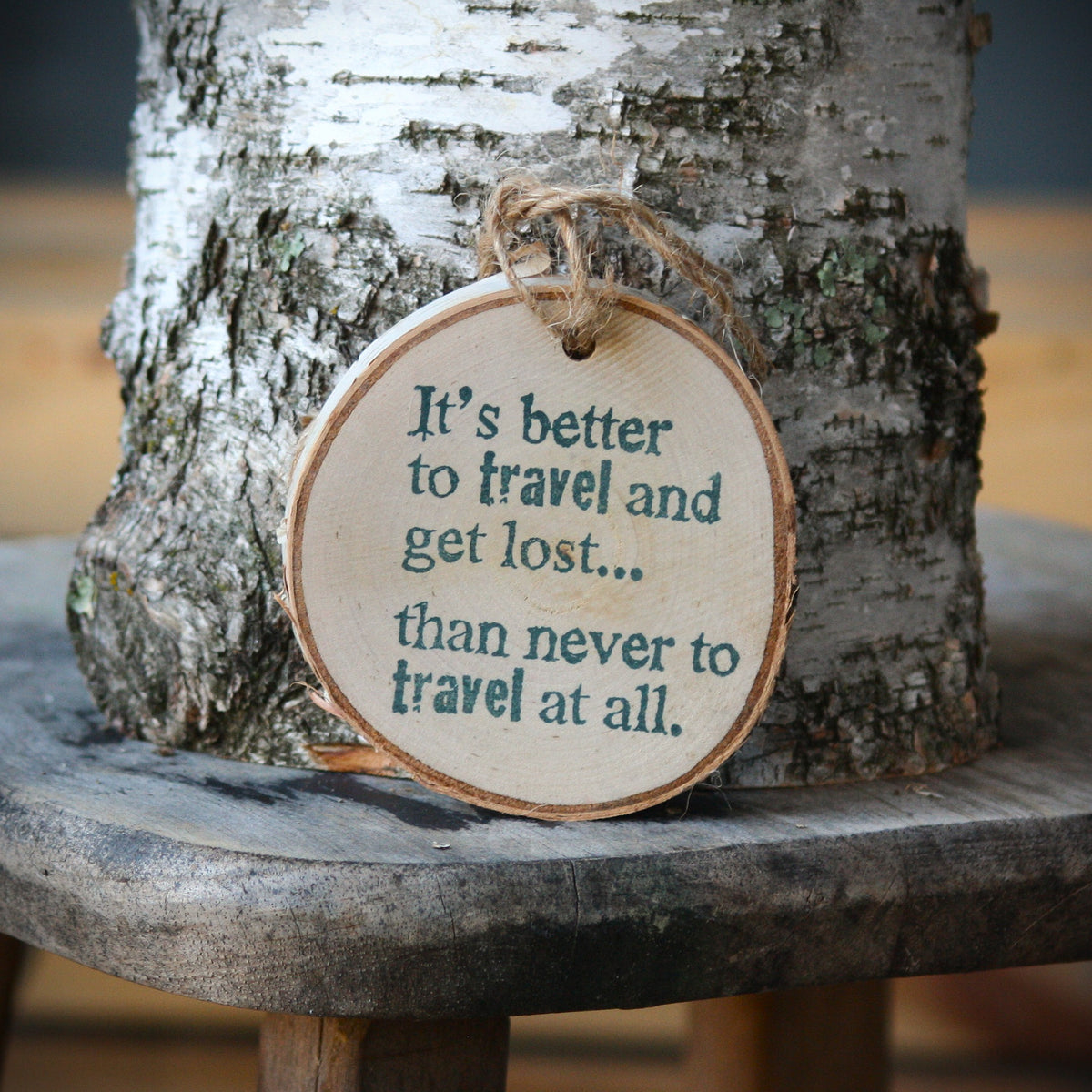 A round wooden hanging ornament with a natural wood edge and twine loop, printed with the quote "It's better to travel and get lost.... than never to travel at all." Made by Log Slices by Margie aka Hayes Street Candle Company of Ferndale, Washington, USA.