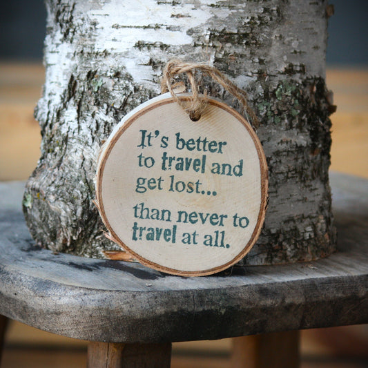 A round wooden hanging ornament with a natural wood edge and twine loop, printed with the quote "It's better to travel and get lost.... than never to travel at all." Made by Log Slices by Margie aka Hayes Street Candle Company of Ferndale, Washington, USA.