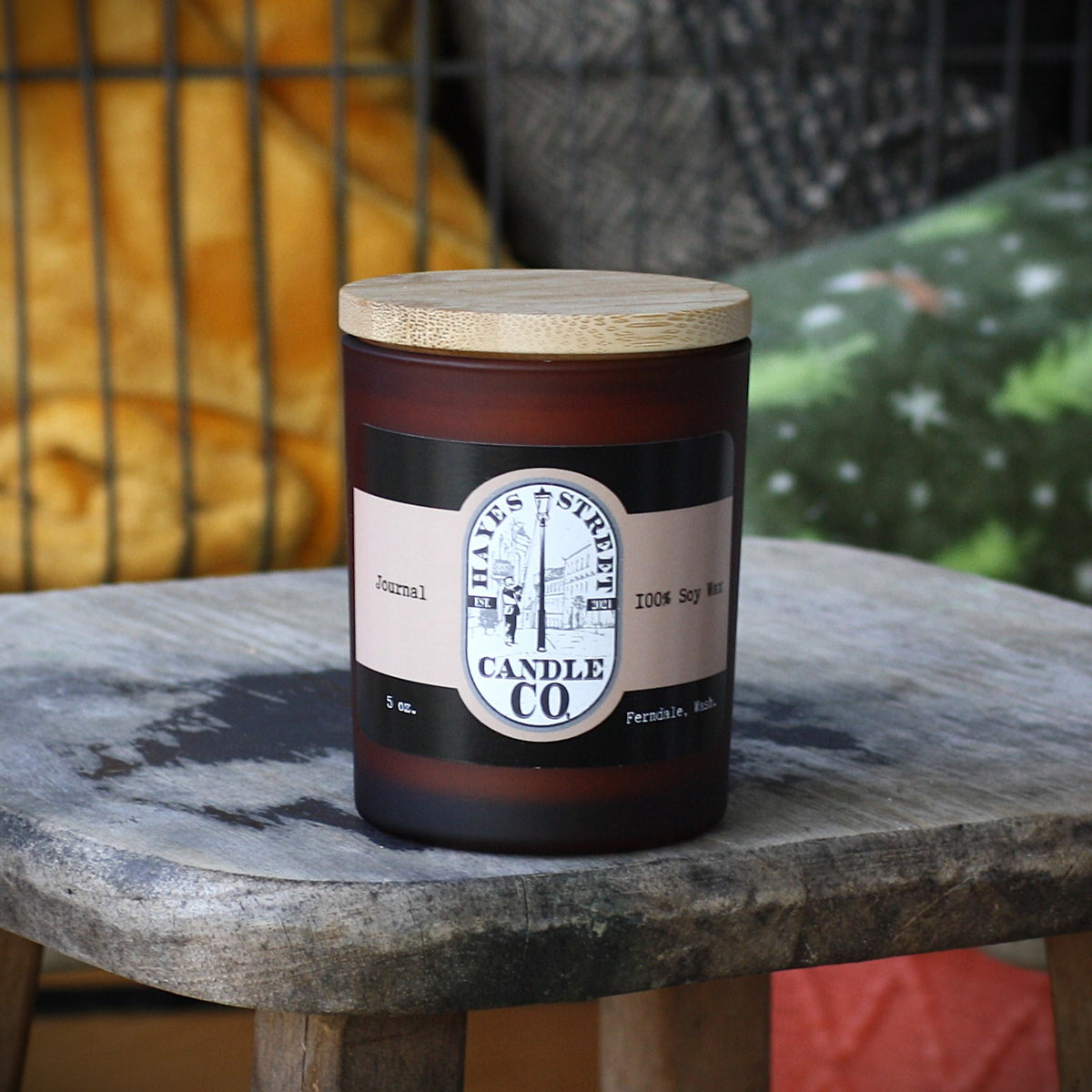 A 5 oz soy candle in a dark amber glass jar with a light wood lid, sitting on a rustic stone table. The label reads 'Hayes Street Candle Co. It also says Ferndale, Wash and Journal