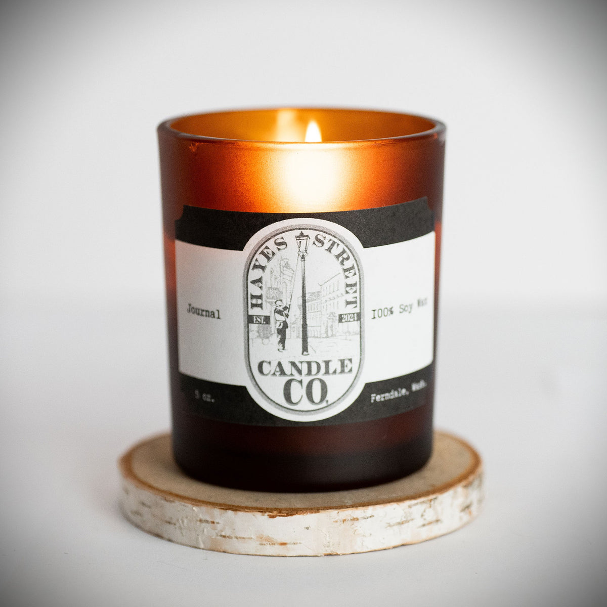 An amber glass jar candle with a lit flame, resting on a round distressed wood coaster. The candle features a white and black paper label for Hayes Street Candle Co. The scent is Journal.