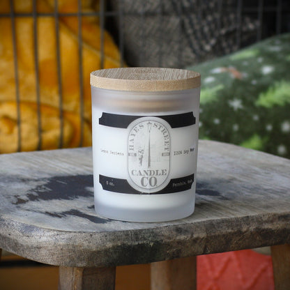 A frosted glass candle jar with a light wood lid sits on a rustic wooden table. The front label reads 'HAYES STREET CANDLE CO' with 'Lemon Verbena' and '5oz Soy Wax' visible.