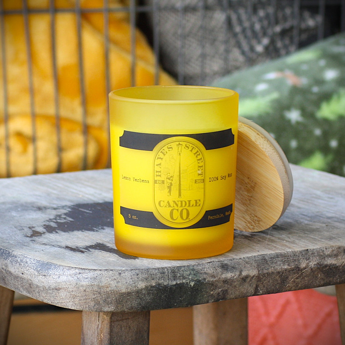 A frosted yellow glass candle jar with a black and gold label reading 'HAYES STREET CANDLE CO.' sits on a rustic wooden table, with its light wood lid resting beside it. Lemon Verbena scent