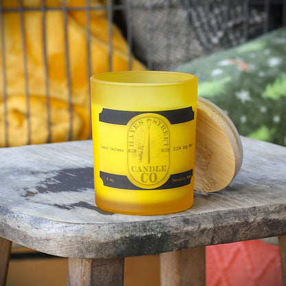 A frosted yellow glass candle jar with a black and gold label reading 'HAYES STREET CANDLE CO.' sits on a rustic wooden table, with its light wood lid resting beside it. Lemon Verbena scent